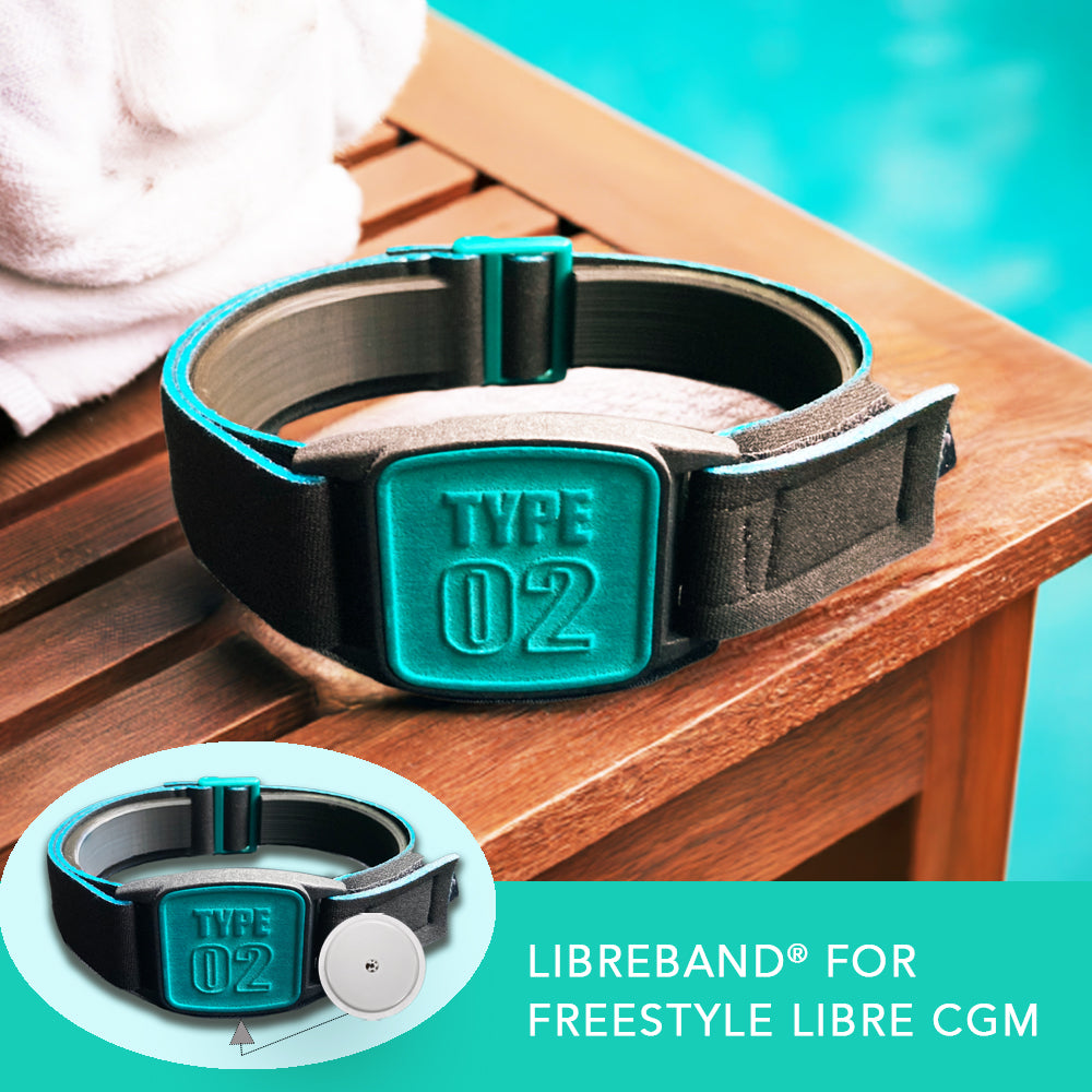 A Freestyle Libre 2 CGM sports and swim armband in teal and black, placed on a wooden surface near a white towel and pool in background.