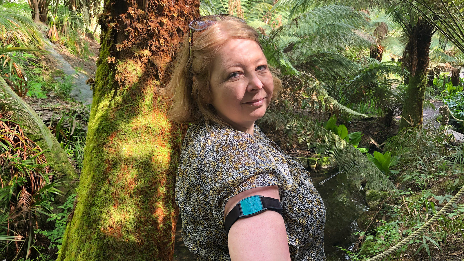 Libreband CGM armband with teal cover worn on upper arm of female in garden setting.